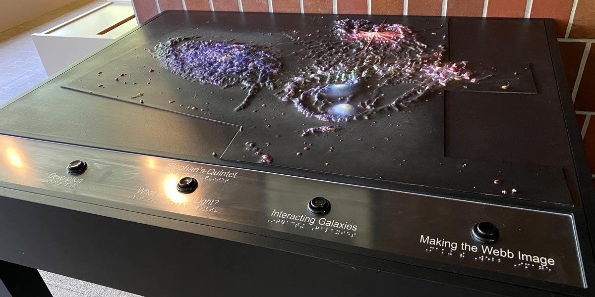 A table with a tactile display of Stephan’s Quintet on top takes up most of the frame. It appears to be sitting in an open room, against a red brick wall. The galaxies vary in terms of texture, elevation of certain characteristics, and colors. The galaxy on the left is mostly dark purple, and the texture resembles puffy, clump-like material. Toward the back of the table, there is splotchy red, vein-like material. Toward the front, there are two smooth areas of light blue and purple material surrounded by a half-circle ridge of bumpy, gray material. There are labeled buttons across the front of the table, but only two can be read. The second from right, “Interacting Galaxies,” and far right, “Making the Webb Image.” Braille runs below each button’s label. 